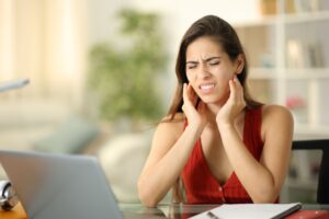 Woman in red shirt at table with laptop rubbing jaw in pain